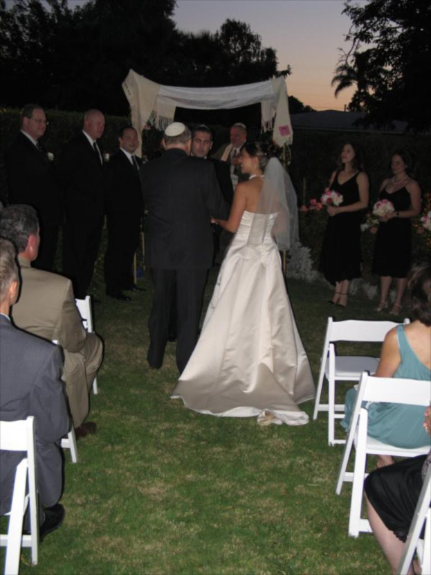 Eine Gruppe von Menschen bei einer Hochzeit, wobei die Frauen auf der rechten Seite Blumensträuße halten, während andere auf Stühlen sitzen und Bäume im Hintergrund zu sehen sind.