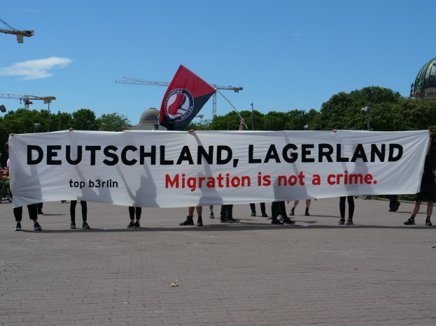 Eine Gruppe von Menschen mit einem Transparent, auf dem 'Deutschland, Lagerland Migration ist kein Verbrechen' steht, und einer Flagge, im Hintergrund sind Laternenpfähle, Straßenlaternen, Bäume, Kräne, ein Gebäude und ein bewölkter Himmel zu sehen.