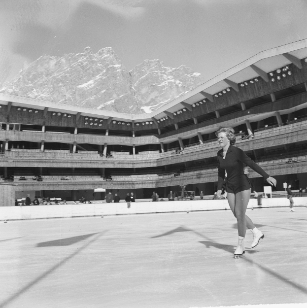 Eine Frau beim Eislaufen auf einer Eisfläche mit Zuschauern in der Tribüne, vor einer Bergkulisse unter einem blauen Himmel.
