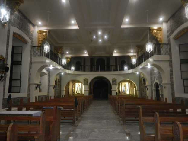 Das Innere einer Kirche mit hölzernen Kirchenbänken, Tischen in der Mitte, Geländern, Lampen und Skulpturen, mit detaillierten Wandmalereien und einer warm beleuchteten Decke.