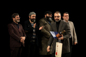 Eine Gruppe von Männern steht auf einer Bühne zusammen, einer hölt eine Tasche und eine Trophäe, mit einem dunklen Hintergrund, der die Eröffnung der Berliner Filmfestspiele suggeriert.