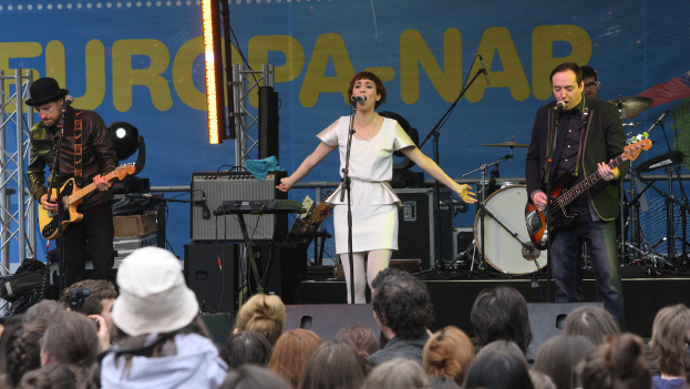 Eine Bühne mit vier Musikern: Eine Frau in einem weißen Kleid singt in der Mitte, zwei Männer spielen Gitarren auf der linken und rechten Seite, eine Menschenmenge vorne und Lautsprecher an der Wand im Hintergrund.