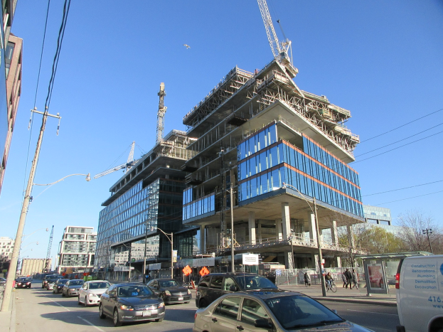 Ein großes Gebäude im Bau auf einer Straßenecke mit Fahrzeugen, Fußgängern, Strommasten, Laternen, Bäumen und einem klaren blauen Himmel.