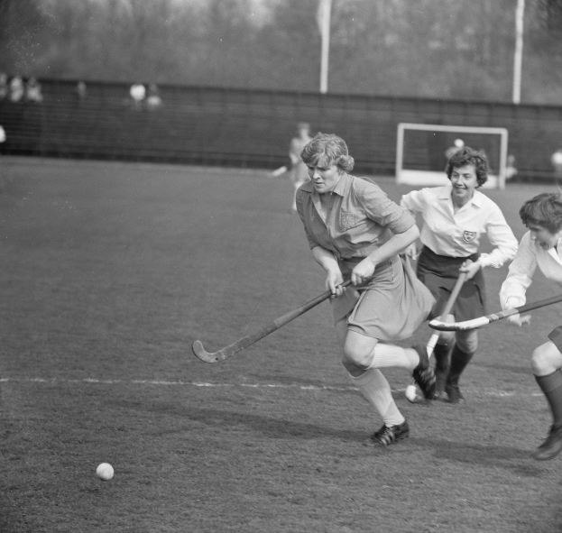 Eine Gruppe von Frauen, die Feldhockey auf einem Feld spielt, mit einem Ball in der Mitte der Aktion, einem Torpfosten, einer Wand, Pfosten, Bäumen und einigen Zuschauern im Hintergrund, dargestellt in Schwarz-Weiß.