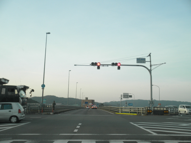 Ein Auto fährt auf einer Autobahn mit einer Ampel an der Kreuzung, Fahrzeuge auf der Straße, eine Person auf dem Gehweg, Geländer, Laternen, Verkehrszeichen, Texttafeln, Gebäude, Hügel und einen klaren blauen Himmel im Hintergrund.