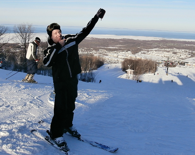 Eine Person auf einem Snowboard in der Mitte des Bildes, die eine Abfahrt mit Bäumen, anderen Menschen, einem Pfahl, Wasser und Himmel im Hintergrund hinunterfährt.