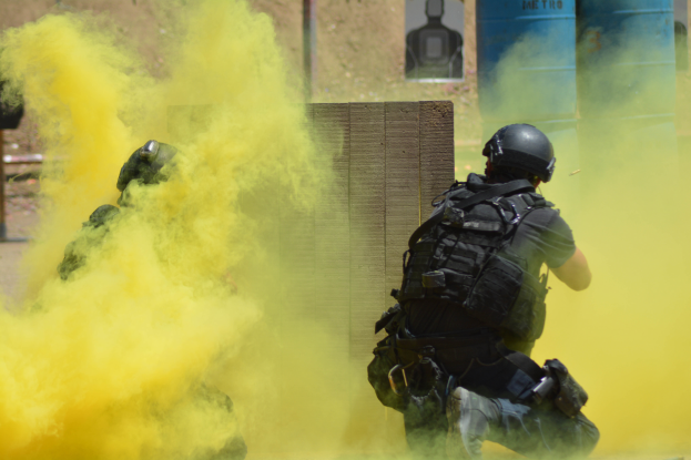 Zwei Polizisten in Einsatzausrüstung mit gelbem Rauch aus dem Mund, die Helme und Schutzausrüstung tragen, stehen vor einer Wand mit einer Tafel und anderen Gegenständen, wobei der Boden unten sichtbar ist.
