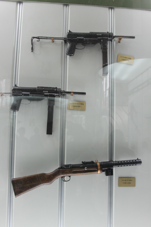 Drei Maschinenpistolen mit langen Läufen und Holzschäften, die in einer durchsichtigen Glasvitrine ausgestellt sind, jeweils mit einer Namensschild