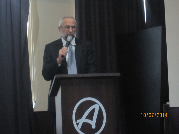 Eine Person in einem schwarzen Mantel und grauen Krawatte steht an einem Schreibtisch und hält ein Mikrofon in der Hand, während sie eine Rede hält, mit schwarzen Vorhängen im Hintergrund.