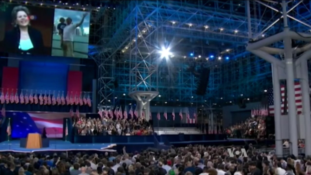 Eine große Menschenmenge sitzt vor einer Bühne mit einem Rednerpult, einem Mikrofon, Flaggen, einem Bildschirm und Beleuchtungsausrüstung und wartet auf eine Präsidentschaftsdiskussion.