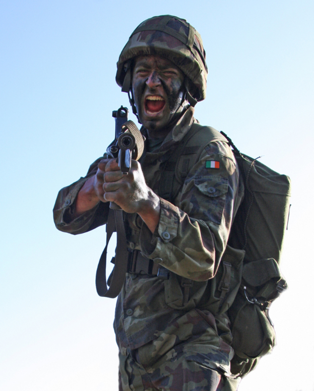 Eine Person in Armeeuniform mit Helm und Tasche hält eine Waffe und trägt ein Abzeichen.