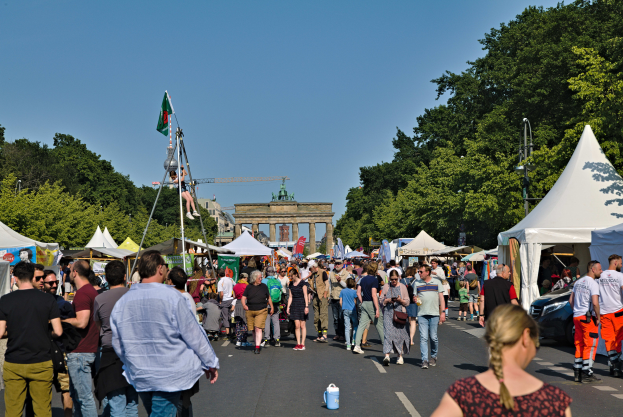 Eine Menschenmenge, die eine Straße mit Zelten, Fahrzeugen und Bäumen entlanggeht, im Hintergrund ein Tor und ein klarer blauer Himmel sowie links Poles mit Fahnen, wahrscheinlich das Oktoberfest in München, Deutschland darstellend.