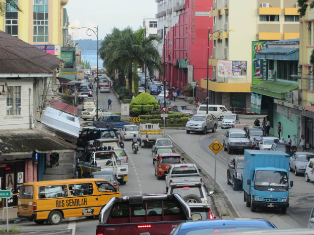 Eine belebte Straße mit Bussen, Fahrrädern und Autos, Menschen auf der rechten Seite und Gebäude, Bäume und Pfosten im Hintergrund.