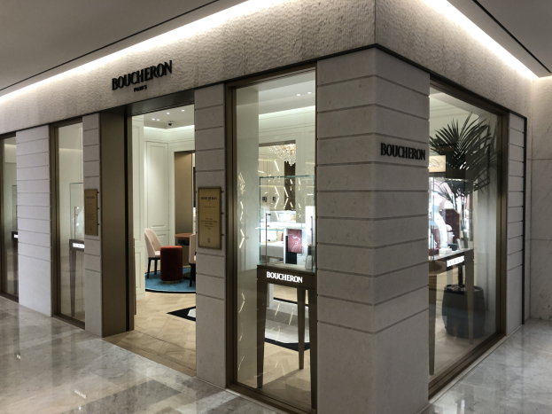 Eingang zu einem Boutique in einem Einkaufszentrum mit Glaswänden und -türen, Text an den Wänden, Deckenleuchten, Stühlen, Tischen, einer Pflanze und anderen Gegenständen, wobei der Boden unten zu sehen ist.