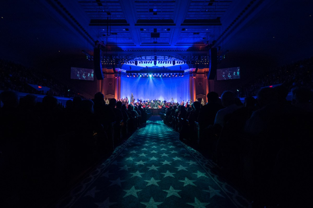 Ein großer Saal mit Reihen von Stühlen, die auf eine Bühne zeigen, auf der Musiker spielen, unter einer Decke mit Lichtern und Fernsehern, was auf einen Konzertsaal während der Nationalen Convention 2019 hinweist.