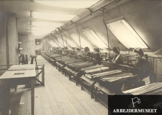 Ein Schwarz-Weiß-Foto einer Gruppe von Frauen, die in einer Textilfabrik an Maschinen arbeiten, die an Tischen auf der linken Seite sitzen, ein Uhr an der Wand im Hintergrund und Text in der rechten unteren Ecke.