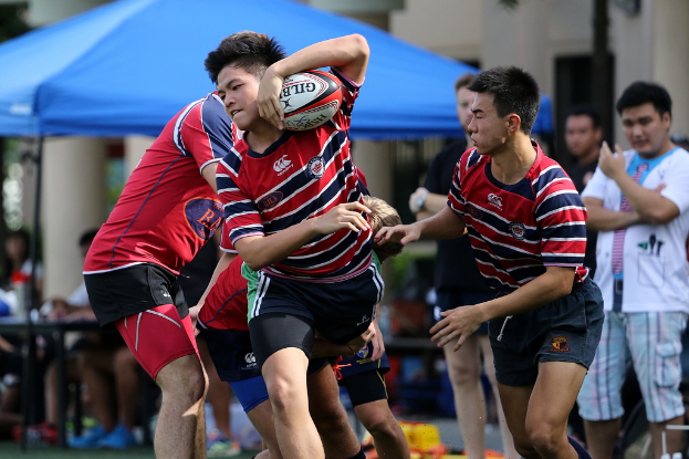 Eine Gruppe von Menschen spielt draußen ein Spiel mit einem Jungen in der Mitte, der den Ball hält, während andere zuschauen und im Hintergrund bei einem blauen Zelt stehen.