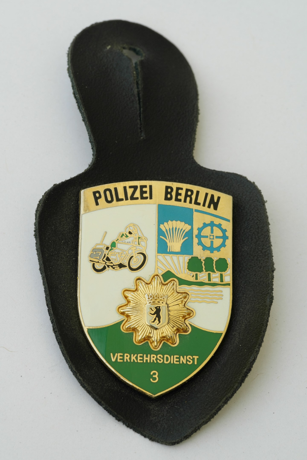 Ein schildförmiges Polizeibadge mit "Polizei Berlin" in schwarzer Schrift auf einem blauen Hintergrund, das ein weißes Stern und einen weißen Rand hat und auf einer Oberfläche liegt.