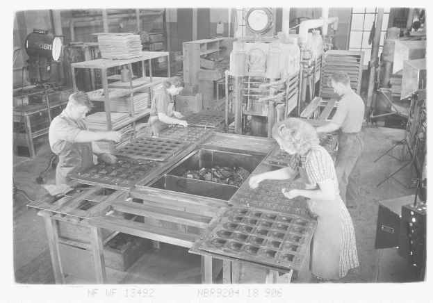 Schwarz-weiß-Bild von Arbeitern, die Essensartikel auf Tischen in einer Fabrikumgebung zusammenbauen, mit Maschinen in der Umgebung.