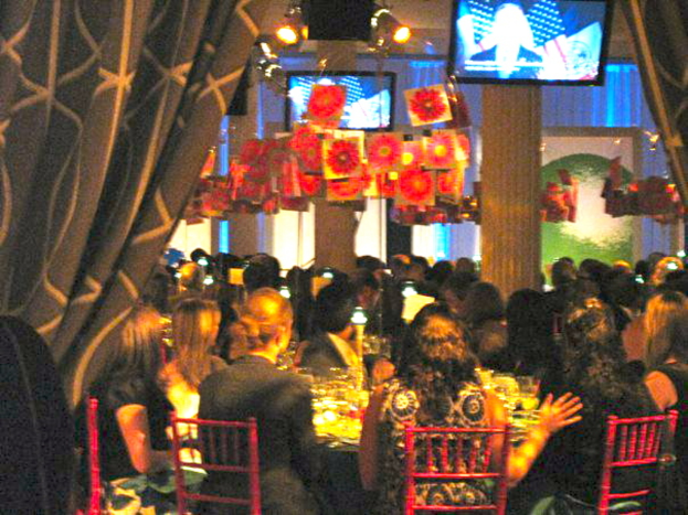 Eine Gruppe von Menschen, die an Tischen mit Essen, Fernsehern, Lampen, hängenden Papieren und Vorhängen in einem Raum sitzt.