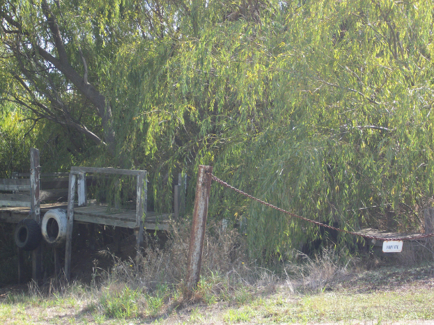 Ein grasbewachsener Bereich mit einem Eisenstab und einer Kette, eine hölzerne Bank links und Bäume im Hintergrund.