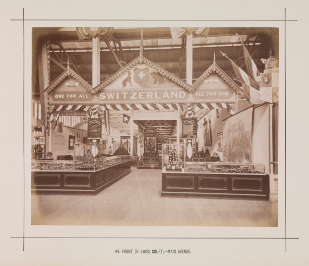 Ein altes Schwarz-Weiß-Foto eines Ladens namens "Schweiz" mit Tischen voller verschiedener Gegenstände, Fotorahmen an den Wänden und Flaggen an der Decke.