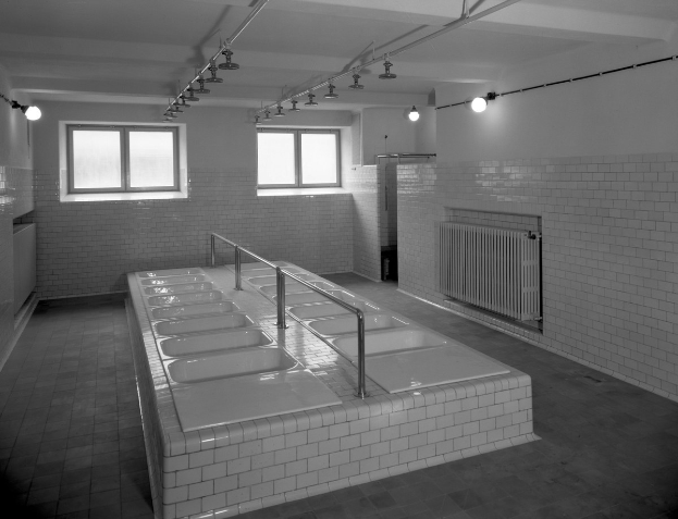 Ein Schwarz-Weiß-Foto einer öffentlichen Toilette mit einer langen Reihe von Waschbecken, gefliesten Wänden, Fenstern, Lampen und sichtbaren Rohren.