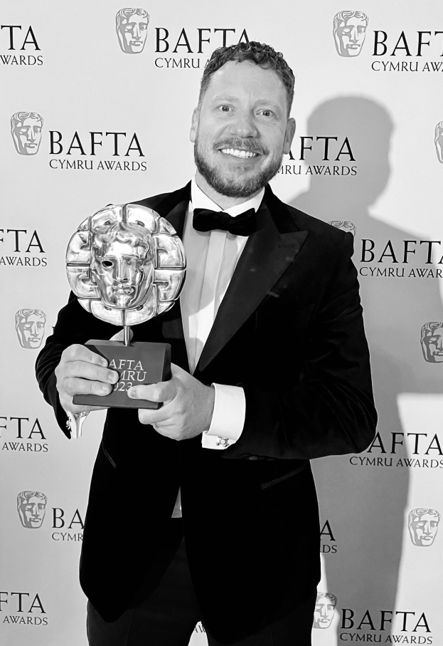 Ein Mann in einem Smoking, der eine BAFTA-Auszeichnung hält und lächelt, mit einem Hintergrund-Schild, auf dem "BAFTA Awards 2018" steht