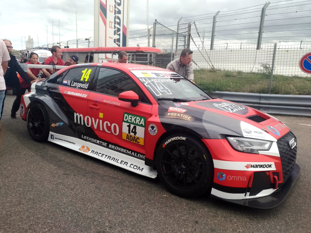 Audi RS3 DTM-Rennwagen auf einer Straße mit einer Gruppe von Menschen, einem Zaun, Gras, einem Schild, einer Fahne, Pfosten und einem bewölkten Himmel im Hintergrund.