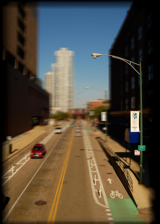 Eine Straßenszene mit Autos auf der Straße, einem Fahrradweg, einer Straßenlaterne mit einem Banner und einem Hinweisschild, Gebäuden und Wolkenkratzern sowie Fahrbahnmarkierungen.
