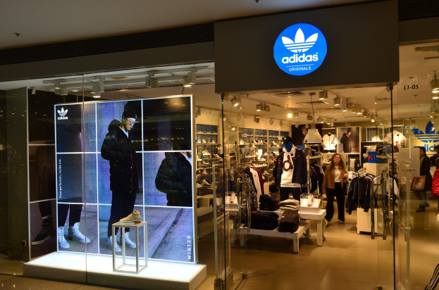 Ein hell erleuchteter Adidas Originals-Store in einem Einkaufszentrum mit Schaufensterpuppen, aufgehängten Kleidern, präsentierten Schuhen und einem Logo-Schild.