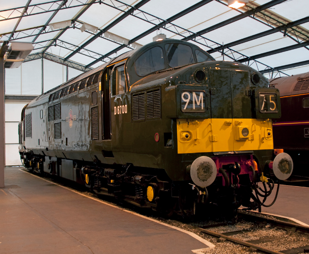 Ein Zug steht auf den Schienen neben einem Bahnsteig mit einem Mast auf dem Dach, der Metallstangen mit Lampen trägt.