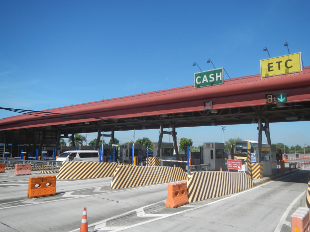 Eine Mautstation mit einem "Barzahlung"-Schild, umgeben von Fahrzeugen, Schranken, Verkehrszeichen, Straßenlaternen, Strommasten, Stromkabeln, Bäumen und einem bewölkten Himmel.