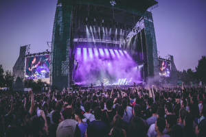 Eine große Menschenmenge bei einem Musikfestival schaut auf der hell erleuchteten Bühne mit Instrumenten und Bildschirmen, die vor einem Gebäude, Bäumen und einem klaren blauen Himmel aufgebaut sind.