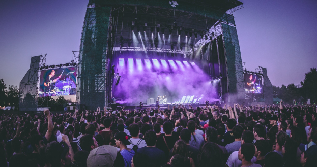 Eine große Menschenmenge bei einem Musikfestival schaut auf der hell erleuchteten Bühne mit Instrumenten und Bildschirmen, die vor einem Gebäude, Bäumen und einem klaren blauen Himmel aufgebaut sind.