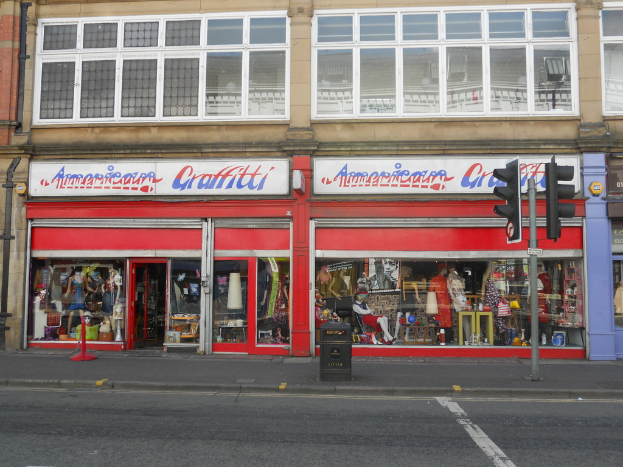 Eine Straßenszene mit einem Gebäude mit Schildern, Puppen, Brillengläsern im Fenster, einem Müllcontainer, einem Pfahl und Verkehrszeichen.